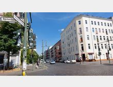 Straßenecke in einer Stadt mit mehrstöckigen Wohngebäuden, darunter ein helles Eckhaus mit zahlreichen Fenstern und kleinen Balkonen. Im Vordergrund stehen Ampeln und Straßenschilder, darunter die Schilder „Reichenberger Straße“ und „Glogauer Straße“, sowie Fußgänger und parkende Autos.