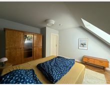 Schlafzimmer mit schräger Decke, einem großen Holzkleiderschrank, einem Doppelbett mit gelber Bettwäsche und blau gemusterten Decken, einem kleinen Holznachttisch, einer Kommode und einem Dachfenster, das Tageslicht einlässt.