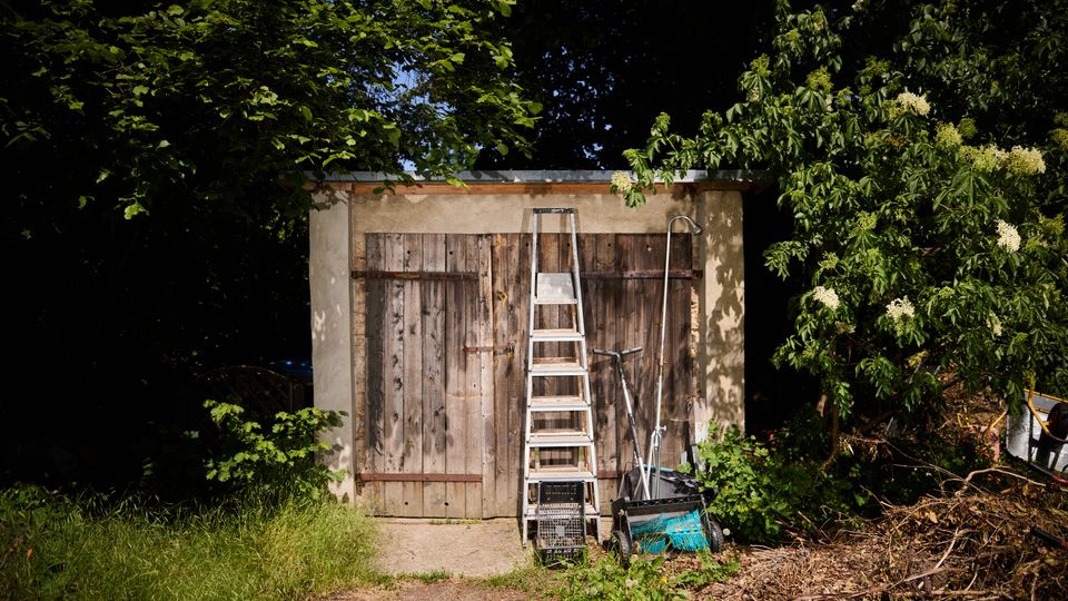 Kleiner Gartenschuppen aus Beton mit zwei verwitterten Holztüren, davor eine angelehnte Leiter und Gartengeräte. Der Schuppen steht in einem von dichtem Grün umgebenen Gartenbereich mit Gras und Gestrüpp im Vordergrund.