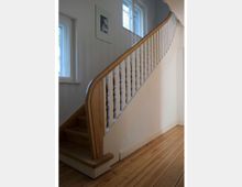 Holztreppe in einem hellen Innenraum mit weiß gestrichenen Wänden, einem Holzgeländer mit weißen Balustern und einem Boden aus Holzdielen. Zwei Fenster mit Rahmen und ein Bild in einem weißen Rahmen befinden sich an der Wand.