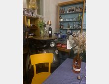 Gemütliche Bar-Ecke in einem Café mit Regalwand voller Spirituosen und Gläser sowie einem Vitrinentresen mit Getränkekisten. Im Vordergrund stehen ein Tisch mit blau-weiß karierter Tischdecke, gelbem Stuhl, Vase mit Blumen und Weinflasche; daneben ist ein Motorroller als Dekoration abgestellt.