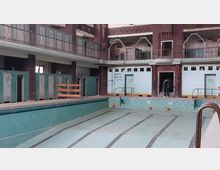 Ein leeres, historisches Hallenbad mit grünlichen Fliesen, Umkleidekabinen und einer umlaufenden Galerie. Im Hintergrund ist eine Tür mit der Aufschrift „Männer Brausen Toiletten“ sowie eine Person am Eingang zu sehen.