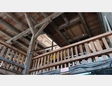 Innenansicht einer Holzkonstruktion mit sichtbaren Balken und Geländern, die Teil eines mehrstöckigen Dachbodens oder Scheunenaufbaus zu sein scheint. Über den Geländern sind Lagergegenstände und lose Kabel erkennbar.