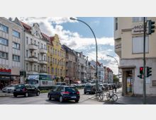 Straßenszene in einem städtischen Viertel mit mehreren mehrstöckigen Wohn- und Geschäftshäusern im historischen Stil, teils mit Balkonen. Auf der Straße befinden sich parkende und fahrende Autos, ein Lkw und zwei Fahrräder an einem Fahrradständer; Ampeln und Verkehrsschilder sind gut sichtbar.