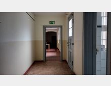 Ein heller Flur mit beige-weißen Wänden und Fliesenboden, der zu einem angrenzenden Raum mit rotem Bodenbelag führt; darüber ein grünes Notausgangsschild. Rechts im Bild eine grau lackierte Tür mit Fenstereinsatz und Blick in einen angrenzenden Raum mit weiß gefliesten Wänden.