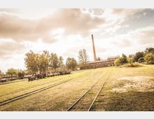 Weitläufiges Industriegelände mit alten Bahnschienen, grasbewachsenen Flächen und vereinzelten Bäumen. Im Hintergrund stehen ein langgestrecktes Backsteingebäude und ein hoher Schornstein, umgeben von einer ländlichen Umgebung.