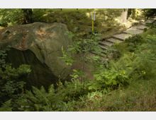 Großer Felsen in einer bewaldeten Parklandschaft, umgeben von Farnen und kleinen Bäumen. Rechts führt ein schmaler, steinerner Treppenweg den Hang hinauf, gesäumt von grasbewachsenen und steinernen Rändern.