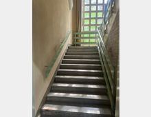 Innen-Treppenhaus mit breiten grauen Steinstufen und beidseitigen hellgrünen Metallgeländern. Am oberen Ende sorgt ein großes Rasterfenster für Tageslicht; rechts eine Ziegelwand, links eine verputzte Wand mit Vorhang.