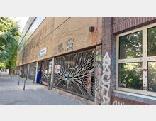 Außenansicht eines Backsteingebäudes mit einem Parkhaus, das durch verzierte Metallgitter mit abstrahierten Baumstrukturen abgedeckt ist. Die Wände sind mit Graffiti bemalt, und im Hintergrund sind Gehwege sowie Bäume zu erkennen.