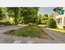 Grüne Innenhofanlage mit Bäumen, Sträuchern und einem kleinen Hügel in der Mitte. Rechts ist ein gepflasterter Gehweg, daneben ein weißes Gebäude mit großen Fenstern sichtbar.