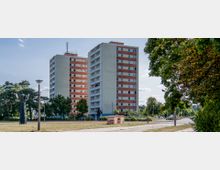 Zwei identische, rechteckige Hochhäuser mit rot-weißer Fassade stehen nebeneinander in einer Grünanlage, umgeben von Bäumen und einem gepflasterten Weg. Im Vordergrund befinden sich eine skulpturähnliche Laterne, weitere Straßenlaternen und ein kleiner Versorgungskasten.