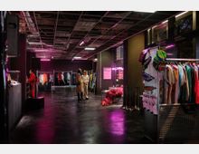 Ein moderner Ladenraum mit industriellem Design, offenem Deckengitter und rosa Beleuchtung. Kleidung hängt auf Metallständern, verschiedene Accessoires sind ausgestellt, und einige Personen stehen und unterhalten sich in der Mitte des Raumes.