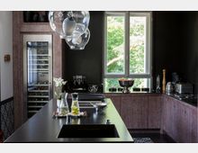 Moderne Küche mit dunklen Wänden, einer Arbeitsplatte aus schwarzem Stein und einer Spüle im Vordergrund. Im Hintergrund stehen ein Weinkühlschrank, eine Kaffeemaschine und eine Schale mit Obst unter einem großen Fenster, das den Blick auf grüne Bäume bietet.