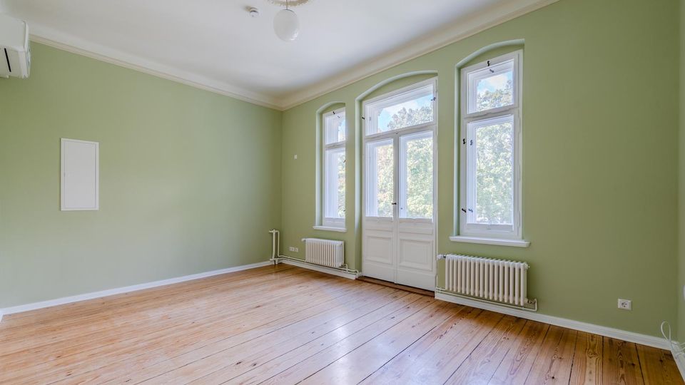 Leerer Wohnraum mit hellgrünen Wänden, hohen Decken mit Stuckrosette und Holzfußboden. Drei hohe Fenster sowie eine verglaste Flügeltür zur Außenseite, darunter Heizkörper; an der Wand ist eine Klimaanlage montiert.