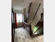 Flur eines Wohnhauses mit hellem Teppichboden, gemustertem Läufer, dunklen Holzvertäfelungen und einer Treppe mit Geländer auf der rechten Seite. Am Ende des Flurs befinden sich eine Tür mit Glaseinsatz und orangefarbene Vorhänge.