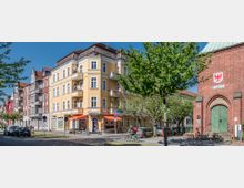 Straßenszene mit einem vierstöckigen gelben Wohngebäude links, das Balkone und eine Pizzeria im Erdgeschoss hat, sowie einem roten Backsteingebäude rechts, das als Amtsgericht gekennzeichnet ist. Im Vordergrund befinden sich Bäume, geparkte Autos, ein Fahrradfahrer und ein Gehweg.