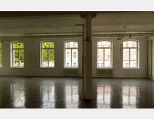 Großer, leerer Raum mit spiegelndem Boden, hohen weißen Wänden und mehreren großen Fenstern mit Blick auf Bäume und eine gegenüberliegende Backsteinfassade. Eine Säule teilt den Raum, und an der Decke sind Lichtschienen montiert.