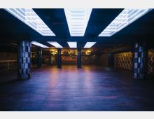 Ein leerer Innenraum mit Holzboden, von rechteckigen, leuchtenden Deckenpaneelen beleuchtet. Säulen und geometrische Wandmuster prägen das moderne Design, und das Licht schafft eine kühle, futuristische Atmosphäre.