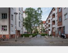 Mehrstöckige Wohnhäuser in einem Innenhof mit weißer und roter Klinkerfassade, flankiert von einem gepflasterten Weg in der Mitte, der von Bäumen und Hecken umgeben ist. Im Vordergrund stehen Laternen, ein Fahrrad und einige Poller.