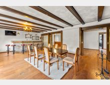 Großer Ess- und Barbereich mit Holzbalkendecke, Holzboden und weiß verputzten Wänden. In der Mitte steht ein Esstisch mit Stühlen auf einem Teppich; im Hintergrund eine gemauerte Theke mit Barhockern, darüber Hängeleuchten und Regale, daneben breite Durchgänge in angrenzende Räume.