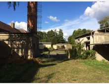 Ein verlassenes Gelände mit mehreren alten Gebäuden, darunter ein Gebäude mit einem Backsteinschornstein und weitere teils verfallene Strukturen. Im Hintergrund sind Bäume und blauer Himmel mit ein paar Wolken zu sehen.