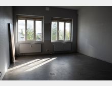 Leerer, grauer Innenraum mit zwei großen Fenstern an der Stirnwand, darunter Heizkörper und sichtbaren Leitungen. Glatte Betonbodenfläche mit Lichtstreifen durch die Fenster und einer an die linke Wand gelehnten Türplatte.