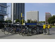 Städtische Szene am U-Bahnhof Ernst-Reuter-Platz in Berlin mit Fahrradständern im Vordergrund und hohen Bürogebäuden im Hintergrund, darunter ein Gebäude mit "Deutsche Bank"-Logo. Gelbe Briefkästen stehen rechts, und mehrere Nationalflaggen wehen auf einem Platz vor den Gebäuden.