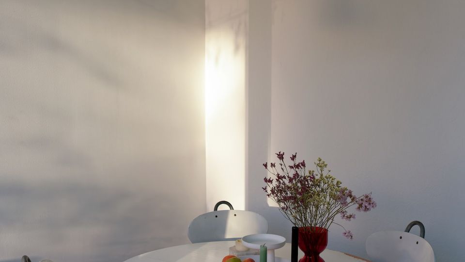 Heller, minimalistischer Essbereich mit rundem weißen Tisch, vier Stühlen mit gepolsterten Sitzkissen und einer weißen Pendelleuchte an hohen, weißen Wänden. Auf dem Tisch stehen eine Obstschale und eine rote Vase mit Blumen, seitliches Tageslicht wirft Schatten an die Wand.