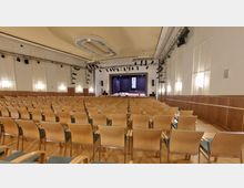 Saal mit einer Bühne im Hintergrund, umgeben von Vorhängen und Beleuchtungstechnik, sowie einer Rednertribüne und einem Flügel. Der Raum ist mit mehreren Reihen hellbrauner Stühle ausgestattet, die sich auf einem Holzfußboden befinden.