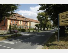 Straßenszene mit einem Ziegelgebäude, das ein Restaurant mit Außenterrasse beherbergt. Im Vordergrund führt eine Straße mit Fußgängerüberweg vorbei, begleitet von Bäumen und einem Wegweiser Richtung „Rosen-Langerwisch, 2000 m“.