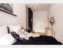 Ein modernes Schlafzimmer mit weiß gestrichenen Wänden und einem Holzboden, ausgestattet mit einem Doppelbett, dekorativen Kissen und einer schwarzen Bettdecke. Im Hintergrund sieht man einen dunklen Akzentbereich, eine Holzgarderobe, eine Kristall-Deckenlampe und minimalistische Möbel.