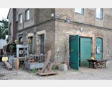 Ecke eines historischen Industriegebäudes aus Backstein mit grünen Holztoren, davor Metallskulpturen, Werkzeuge und abgesperrte Bereiche; auf dem Kopfsteinpflaster steht ein Schild mit „Zutritt verboten“.