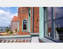 Fassade eines historischen Backsteingebäudes mit grünen Fensterrahmen und dekorativen Elementen, im Hintergrund ein Fluss und angelegte grüne Flächen. Ein Balkon mit weißem Geländer ist im Vordergrund sichtbar.