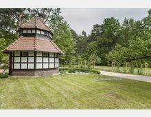 Fachwerkpavillon mit achteckigem Grundriss und rotem Ziegeldach, umgeben von einer Rasenfläche und Bäumen in einem parkähnlichen Umfeld. Im Hintergrund stehen junge, frisch gestützte Bäume entlang eines Gehwegs.