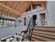 Innenraum eines modernen Hauses mit Holzbalkendecke und offenen Wohnbereichen. Eine Holztreppe mit geschwungenem schwarzen Geländer führt zu einem oberen Geschoss mit weißen Wänden, sichtbaren Holzelementen und mehreren Türen.