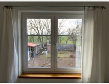 Fensteransicht von innen mit weißen Vorhängen, Blick auf einen Garten mit Hühnern, einer Scheune mit rotem Dach, Bäumen und offener Landschaft im Hintergrund.