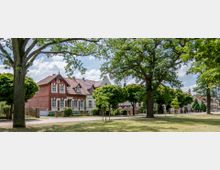 Straßenansicht mit historischen Backsteinhäusern in einer ländlichen Umgebung, umgeben von großen Bäumen und gepflegten Rasenflächen. Der Himmel ist leicht bewölkt, und die Szene wirkt ruhig und grün.