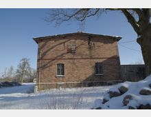 Zweistöckiges Backsteingebäude mit flachem Satteldach und vergitterten Fenstern in einer verschneiten Landschaft. Im Vordergrund liegen Schnee und Steine, daneben ein großer Baum; rechts schließt ein Holzzaun an.