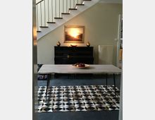 Essbereich mit modernem Tisch auf einem schwarz-weiß gemusterten Teppich vor einer Treppe mit Holzstufen und Geländer. An der Wand hängt ein Bild mit Landschaftsmotiv über einem schwarzen Klavier.