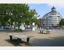 Öffentlicher Platz mit gepflastertem Boden, mehreren Bäumen und zwei Tischtennisplatten im Vordergrund; im Hintergrund ein modernes Wohngebäude mit runder Kuppel, Geschäften im Erdgeschoss und einer Ampelkreuzung.