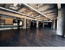 Großer, industriell gestalteter Innenraum mit freiliegenden Betonwänden, Lüftungsrohren an der Decke und dunklem Bodenbelag; im Hintergrund eine moderne Theke mit Holzelementen und schwarz abgesetzten Bereichen, flankiert von Vorhängen.