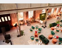 Industrieller Veranstaltungsraum mit Backsteinwänden und hohen Stahlträgern, ausgestattet mit grünen und orangefarbenen Sesseln, kleinen Tischen mit Blumenarrangements sowie Präsentationsflächen mit Postern auf Staffeleien. Durch große rustikale Fenster fällt Tageslicht, ergänzt durch warme Innenbeleuchtung.