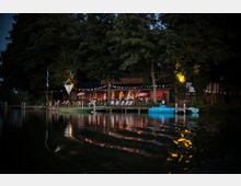 Abendliche Szene an einem Ufer mit beleuchtetem Restaurant, umgeben von großen Bäumen und dekoriert mit Wimpelketten. Im Vordergrund sind ein Holzsteg, Liegestühle, kleine Boote und deren Spiegelungen im Wasser zu sehen.