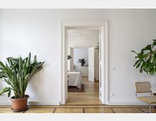 Helle Innenansicht einer Wohnung mit weiß gestrichenen Wänden, einem Parkettboden und großen Zimmerpflanzen. Im Vordergrund ist eine geöffnete Tür zu sehen, die in einen weiteren Raum mit Teppich, Tisch und Kommode führt.