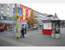 Städtischer Platz mit einem U-Bahn-Eingangsschild der Linie U8, umgeben von gepflastertem Boden, einer öffentlichen Toilette, einem geschlossenen Kiosk und Werbesäulen. Im Hintergrund sind bunte Gebäudefassaden, ein Geschäft und geparkte Fahrzeuge zu sehen.