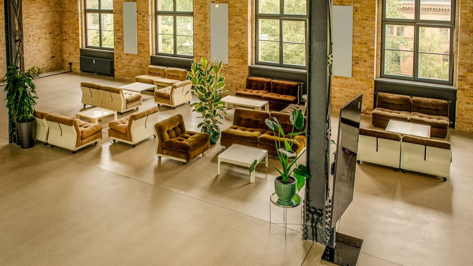 Großer Loft-Loungebereich in einem Industriebau mit freiliegenden Backsteinwänden, Stahlträgern und hohen Sprossenfenstern. Mehrere Sofagruppen mit niedrigen weißen Tischen stehen auf glattem Betonboden, ergänzt durch große Topfpflanzen und eine frei stehende Trennwand.