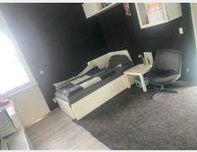 Schlafzimmer mit weißem Tagesbett und grauer Bettwäsche vor dunkel verkleideten Wänden, daneben ein kleiner weißer Beistelltisch und ein grauer Drehsessel. Links fällt Tageslicht durch ein Fenster ein; der Boden ist teils mit hellem Holz und teils mit einem großen grauen Teppich ausgelegt.