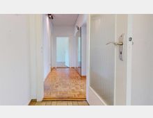 Ein Innenraumflur mit hellem Parkettboden, weißen Wänden und mehreren Türen, die in andere Räume führen. Im Vordergrund ist eine geöffnete Tür mit einer Glasfüllung und silbernem Türgriff zu sehen.