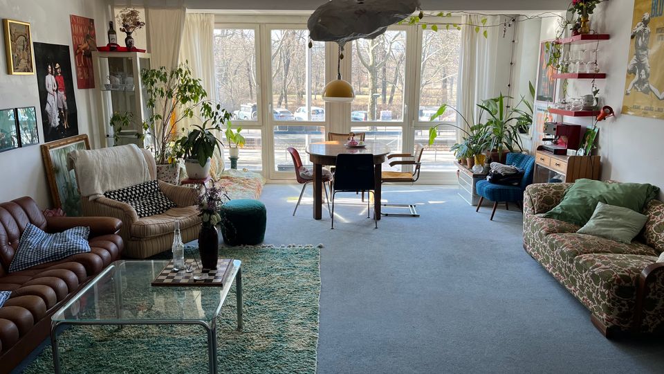 Bright, eclectic living room with large floor-to-ceiling windows and sheer curtains overlooking a street and trees. The space includes mixed vintage seating, a glass coffee table on a rug, a central dining table, wall art and shelves, and many potted plants throughout.