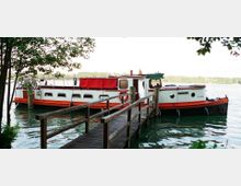 Ein weiß-rotes Hausboot mit schwarzem Unterbau, angedockt an einem hölzernen Steg auf einem ruhigen See. Im Hintergrund sind Bäume und Uferlinien zu sehen.